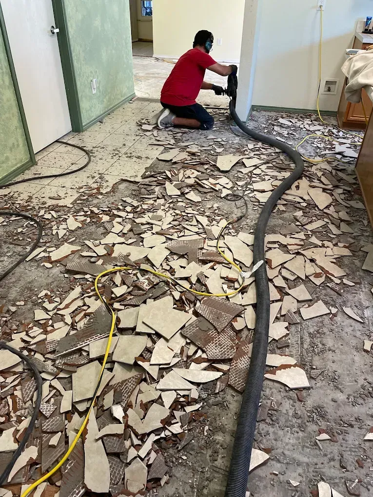 Person in red shirt using demolition tool on floor with tile debris.