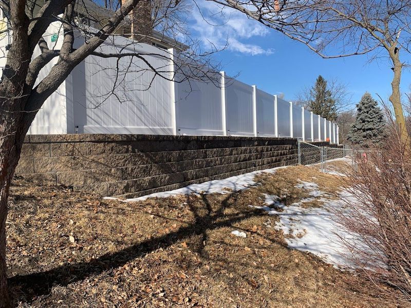 White privacy fence atop a dark retaining wall, on a slightly snowy hillside. Bright sunny day.