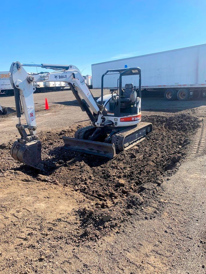 A white Bobcat excavator digs a trench in a dirt area, with a truck trailer visible in the background.