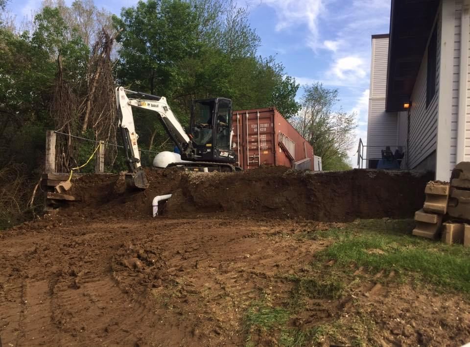 An excavator digs into a dirt slope near a house, with a shipping container visible.