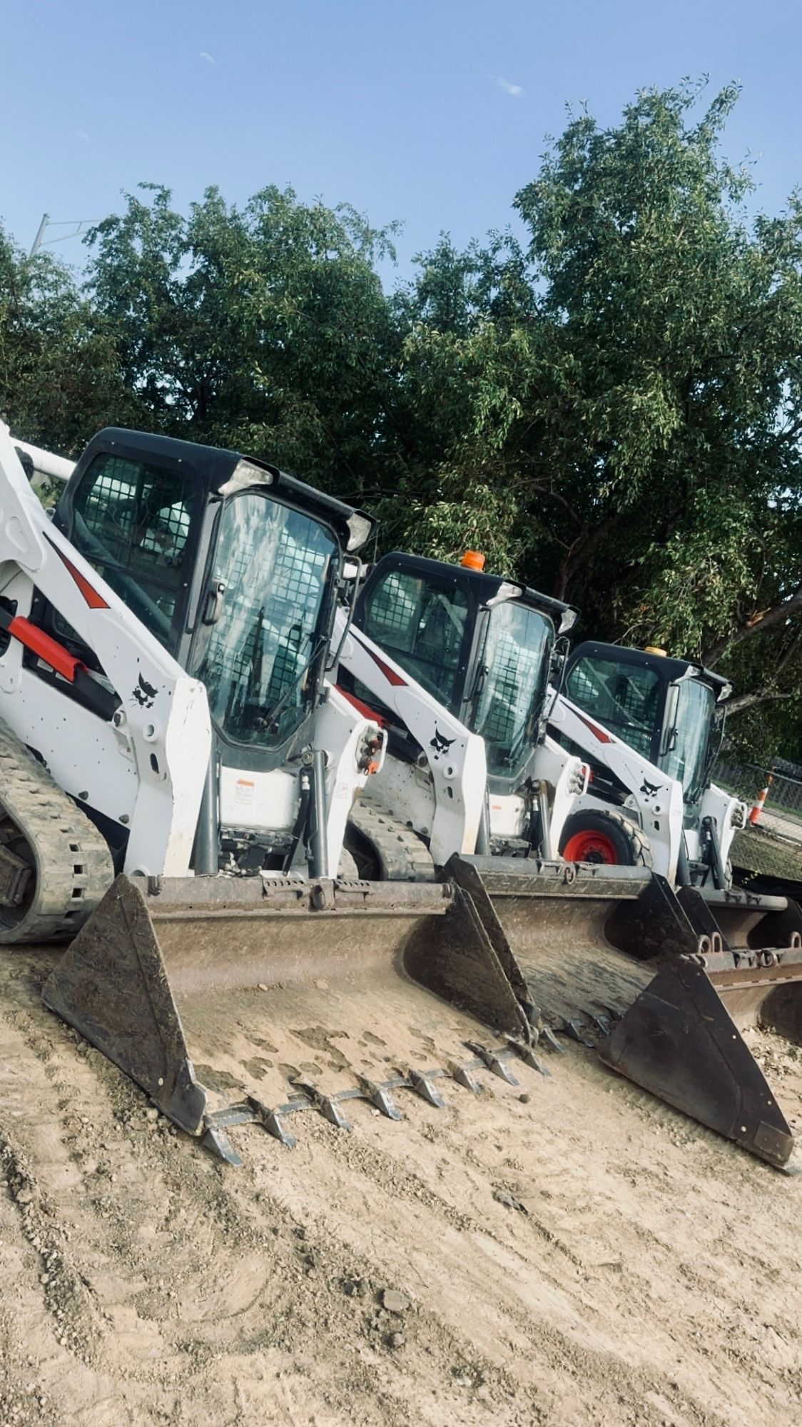Four white Bobcat skid-steer loaders with attached rakes, parked on a dirt surface. Trees are visible in the background.