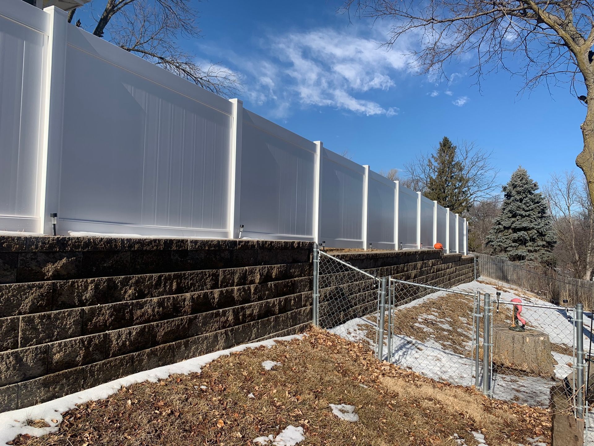 White vinyl fence atop a dark retaining wall on a snowy hillside, under a blue sky.