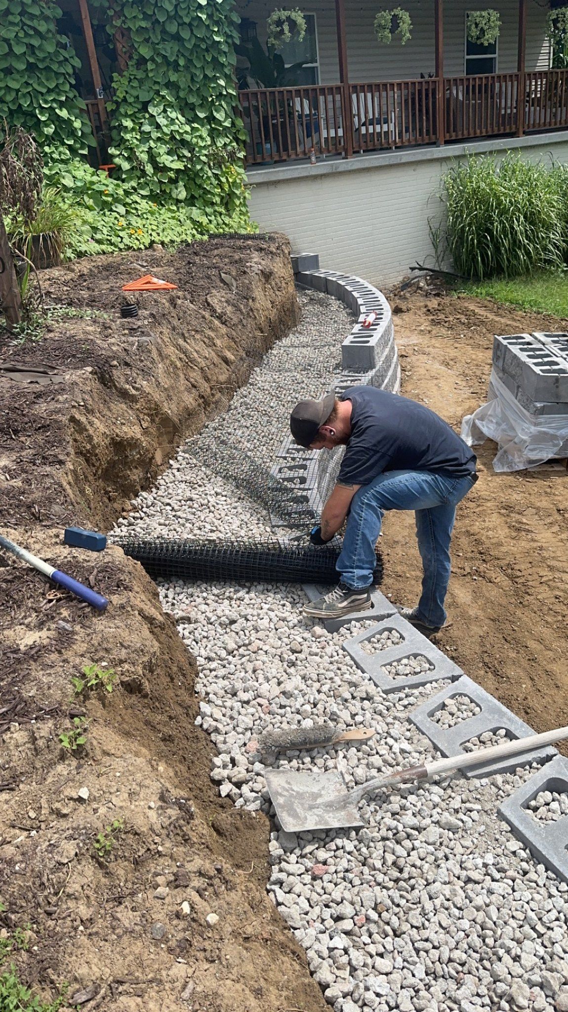 Man building a retaining wall in a yard, gravel and blocks visible, with lush greenery nearby.