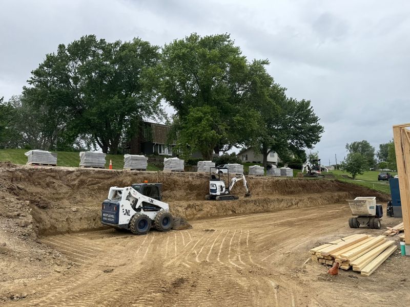 Construction site with a bobcat, excavator, and stacked pavers, next to a house under an overcast sky.