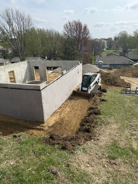 A skid steer excavator excavates dirt next to a concrete foundation for a building under construction.
