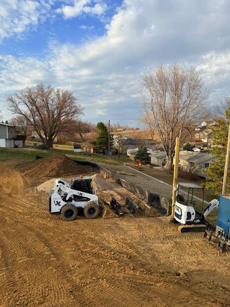 Construction site with Bobcat and mini excavator, grading dirt near houses and trees under a cloudy sky.