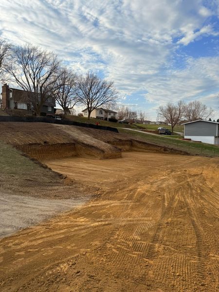 Dirt lot being graded, with homes and trees in the background under a cloudy blue sky.