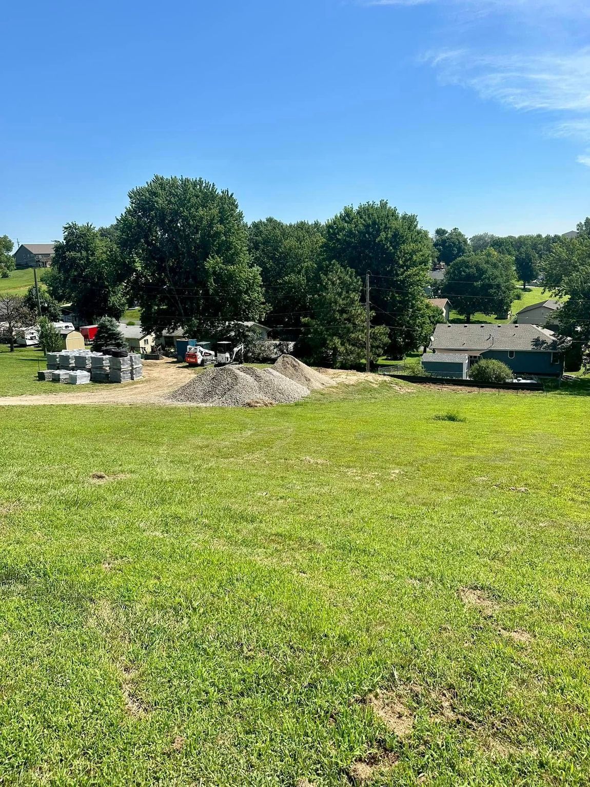 Grassy field with construction materials in the middle distance; trees and houses in the background on a sunny day.