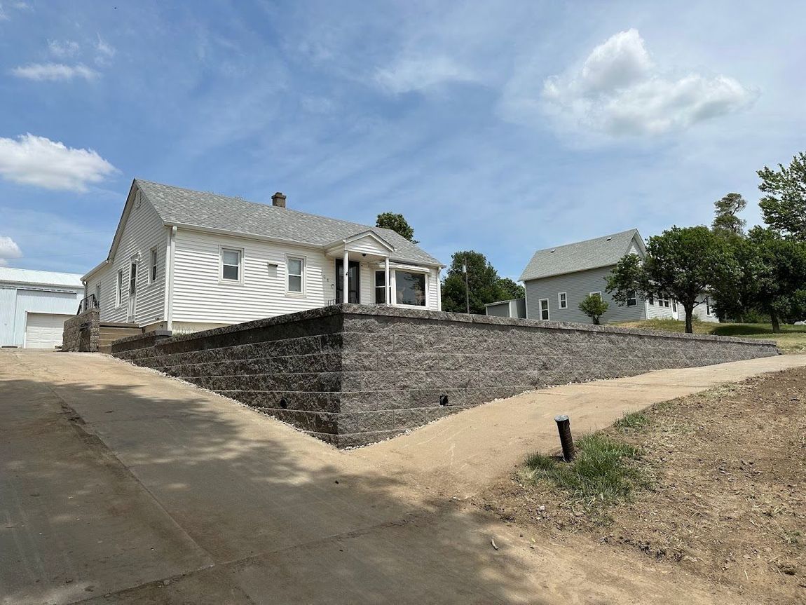 White house and small building with a stone retaining wall, dirt driveway, and blue sky.