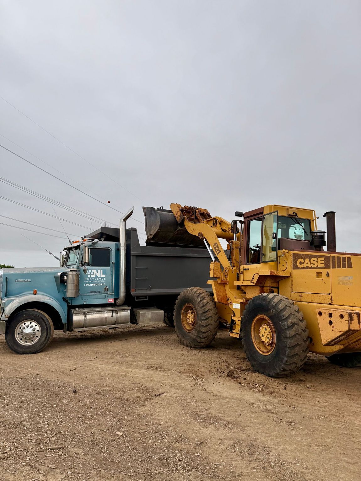 Yellow Case loader dumping dirt into a blue dump truck on a dirt road under an overcast sky.