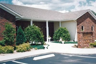 Brick building with arched entrance, columns, and shrubbery. Dark asphalt in foreground, gray roof.