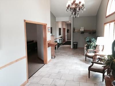 Hallway with chairs, plants, and a chandelier; light-colored walls and flooring.