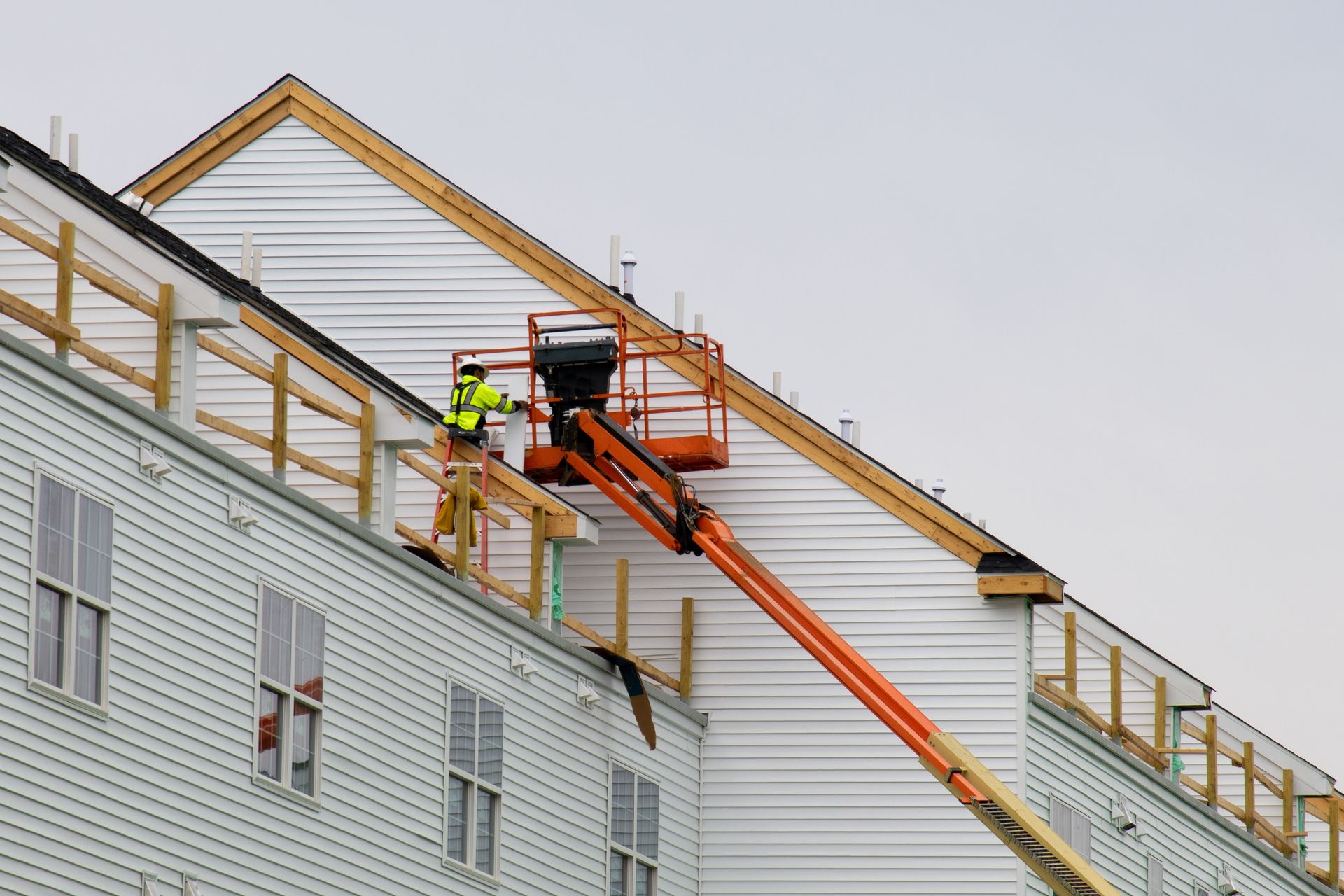 Construction worker on an orange lift works on the exterior of a building with white siding, under a cloudy sky.
