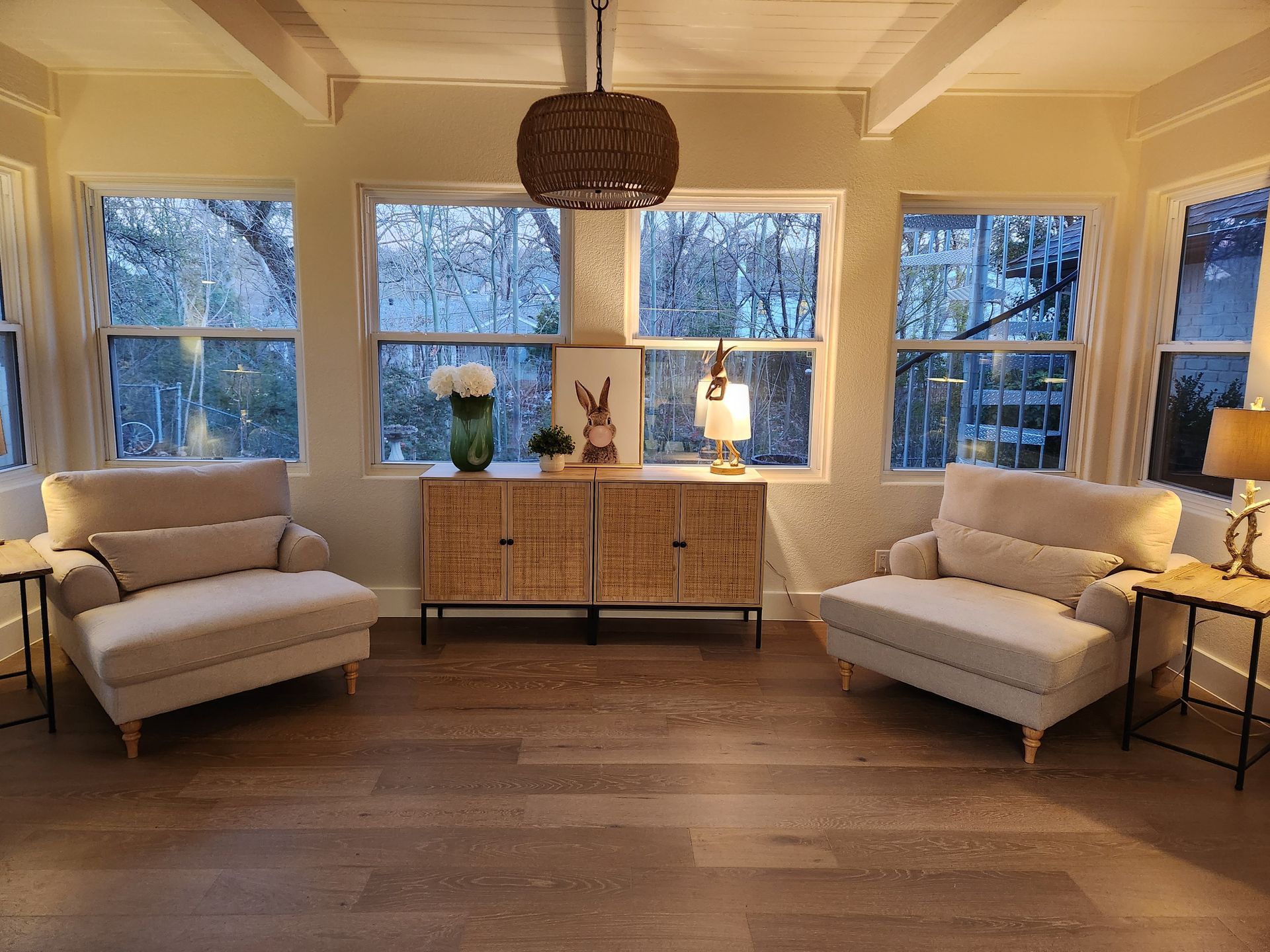 Cozy sunroom with two armchairs, a wooden cabinet, and large windows overlooking a forested area.