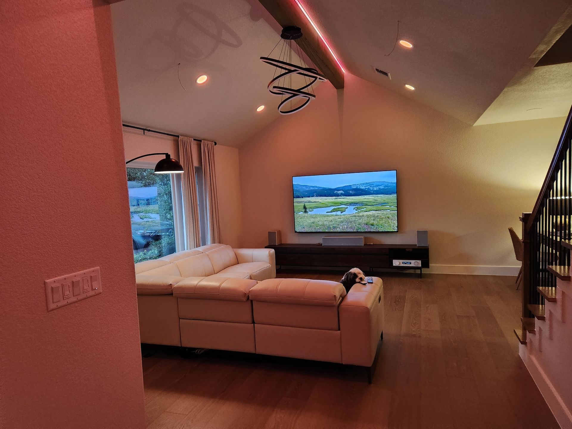 Living room with a large TV above a dark wood console. Cream sectional, hardwood floor, and stairs.