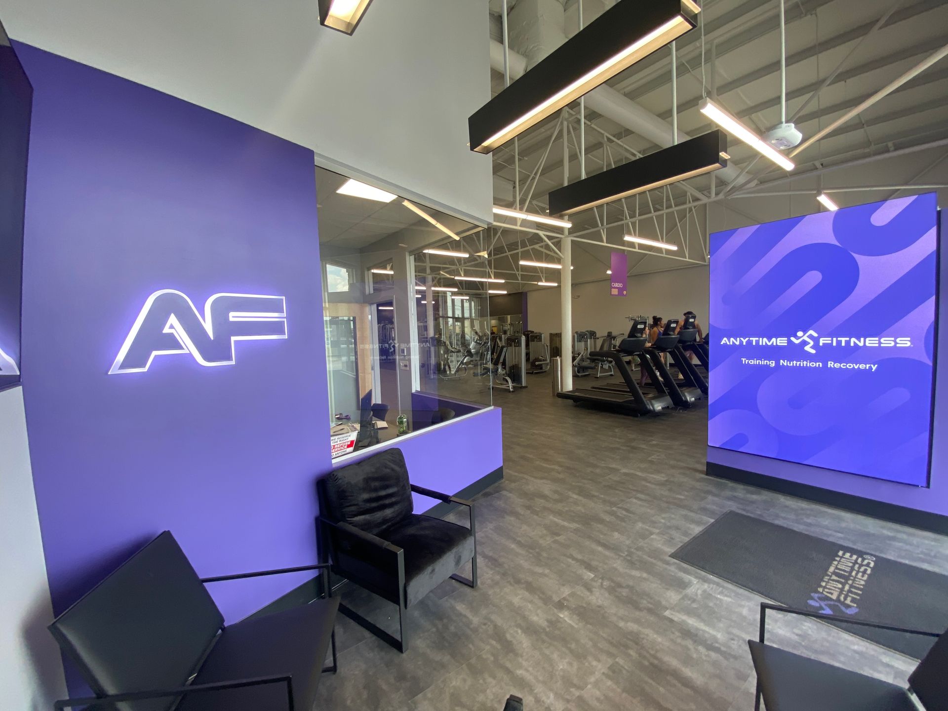 Interior of Active Fitness gym, purple walls, logo, waiting area with black chairs, treadmills visible in the background.