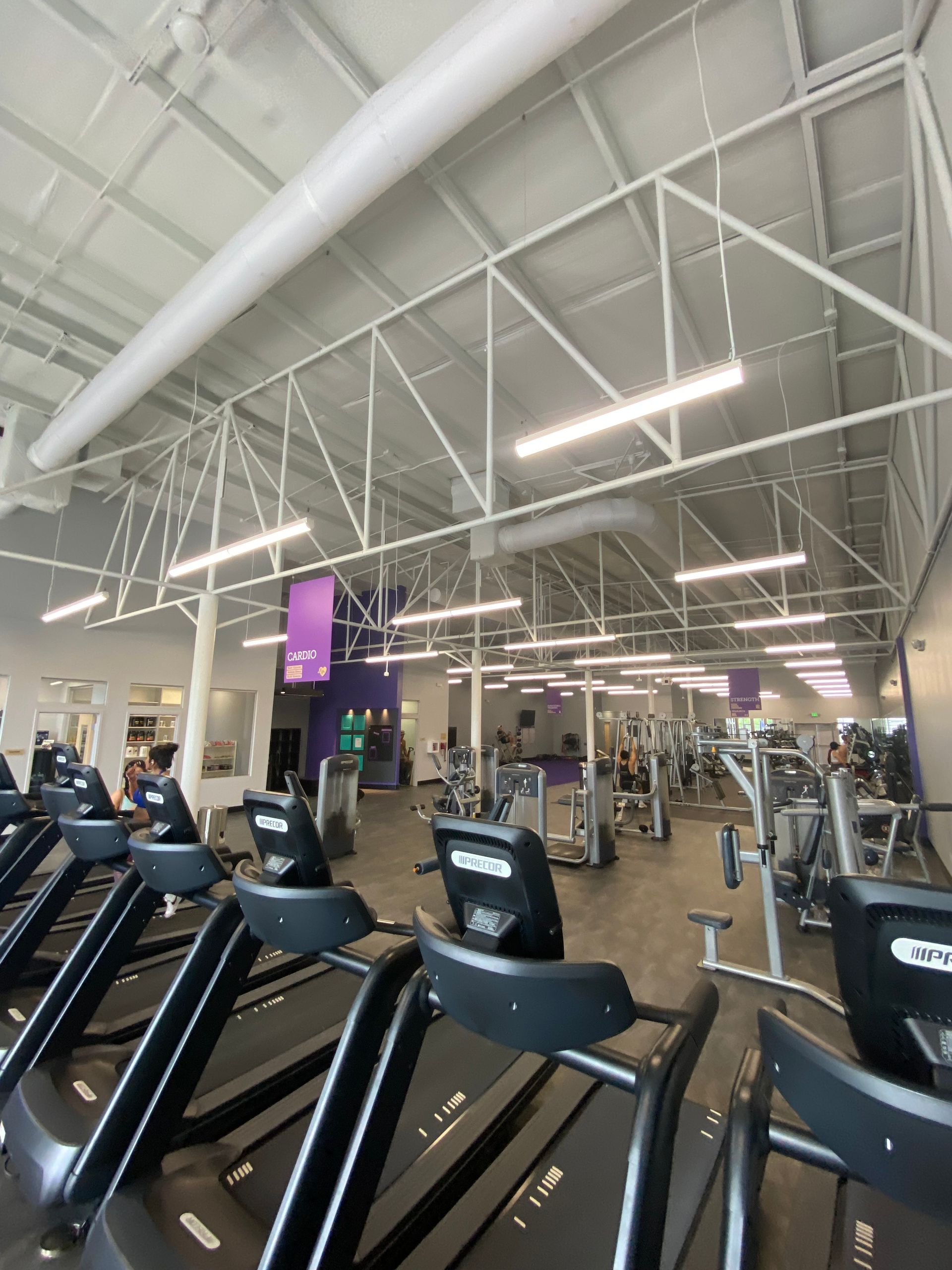 Treadmills and exercise machines fill a brightly lit gym. People work out under a white ceiling and beams.