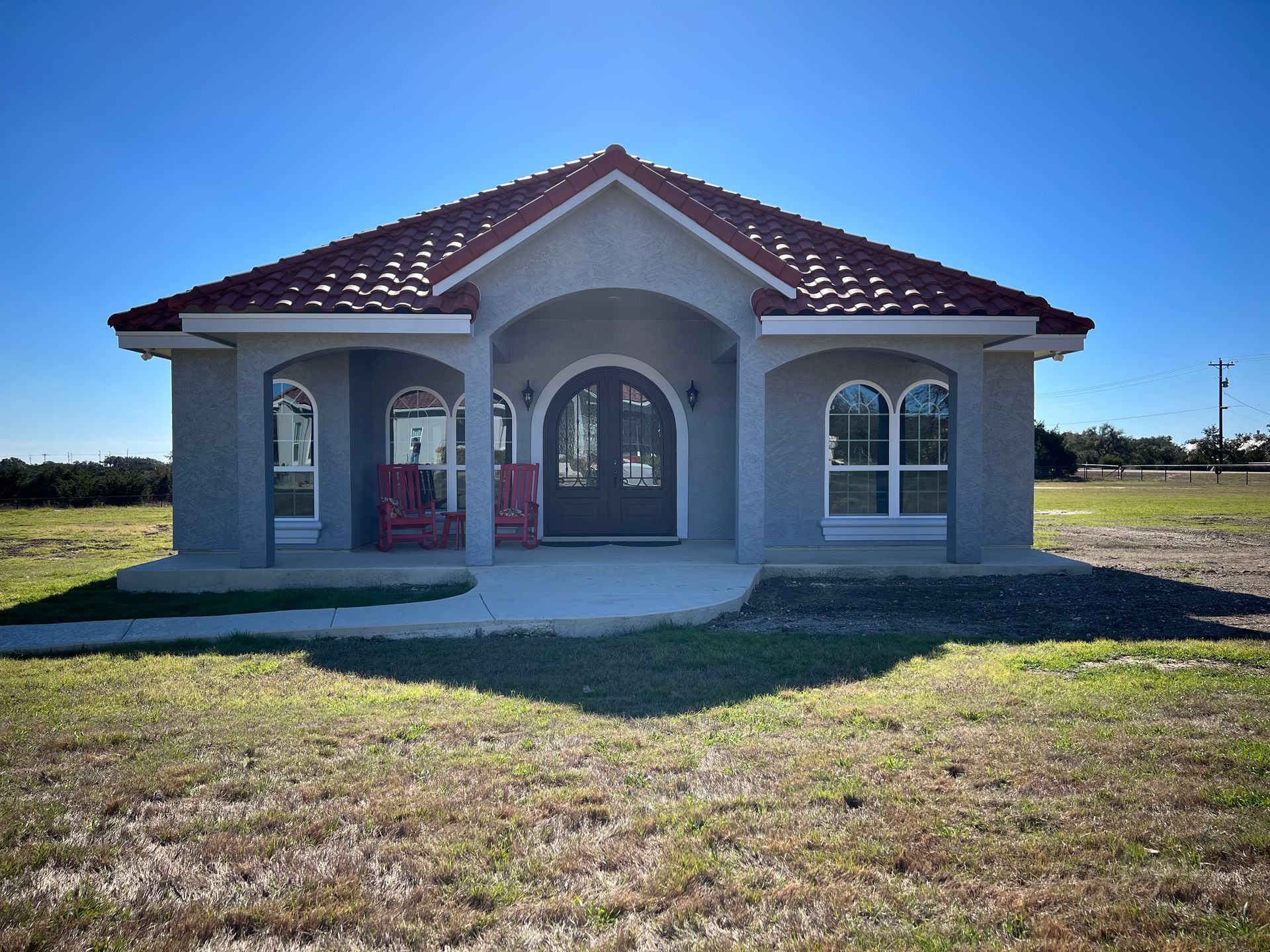 Small stucco house with red-tiled roof; arched windows and doorway; blue sky; grassy field.