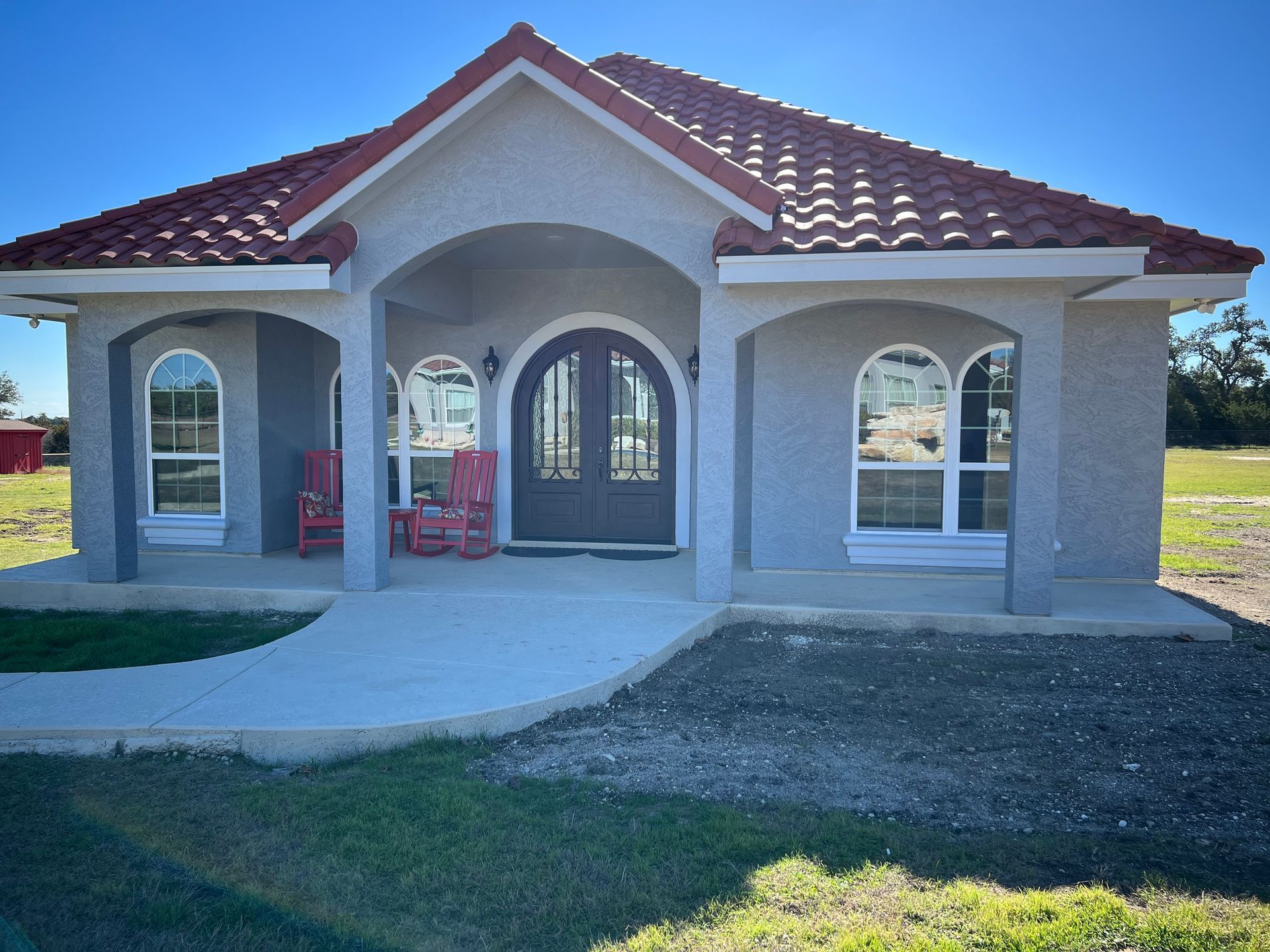 Gray stucco house with red tile roof, arched doorway, windows, and a concrete path.