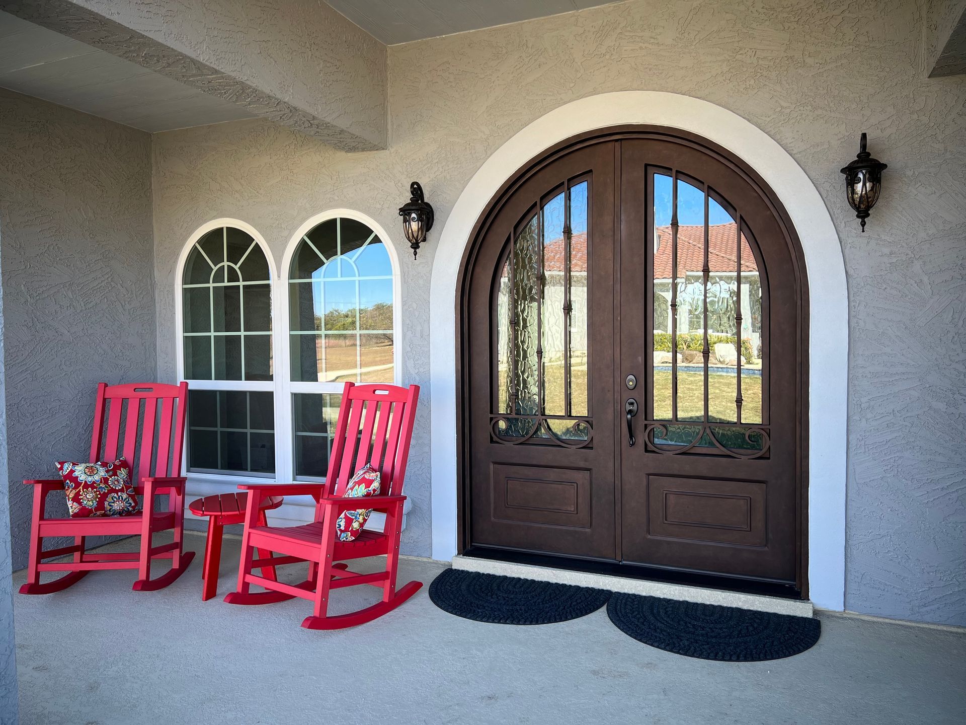 A home's front porch featuring red rocking chairs and arched brown doors with a white border.