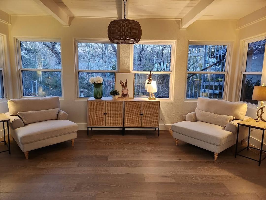 Cozy sunroom with two cream armchairs, a wooden cabinet, and large windows overlooking trees.