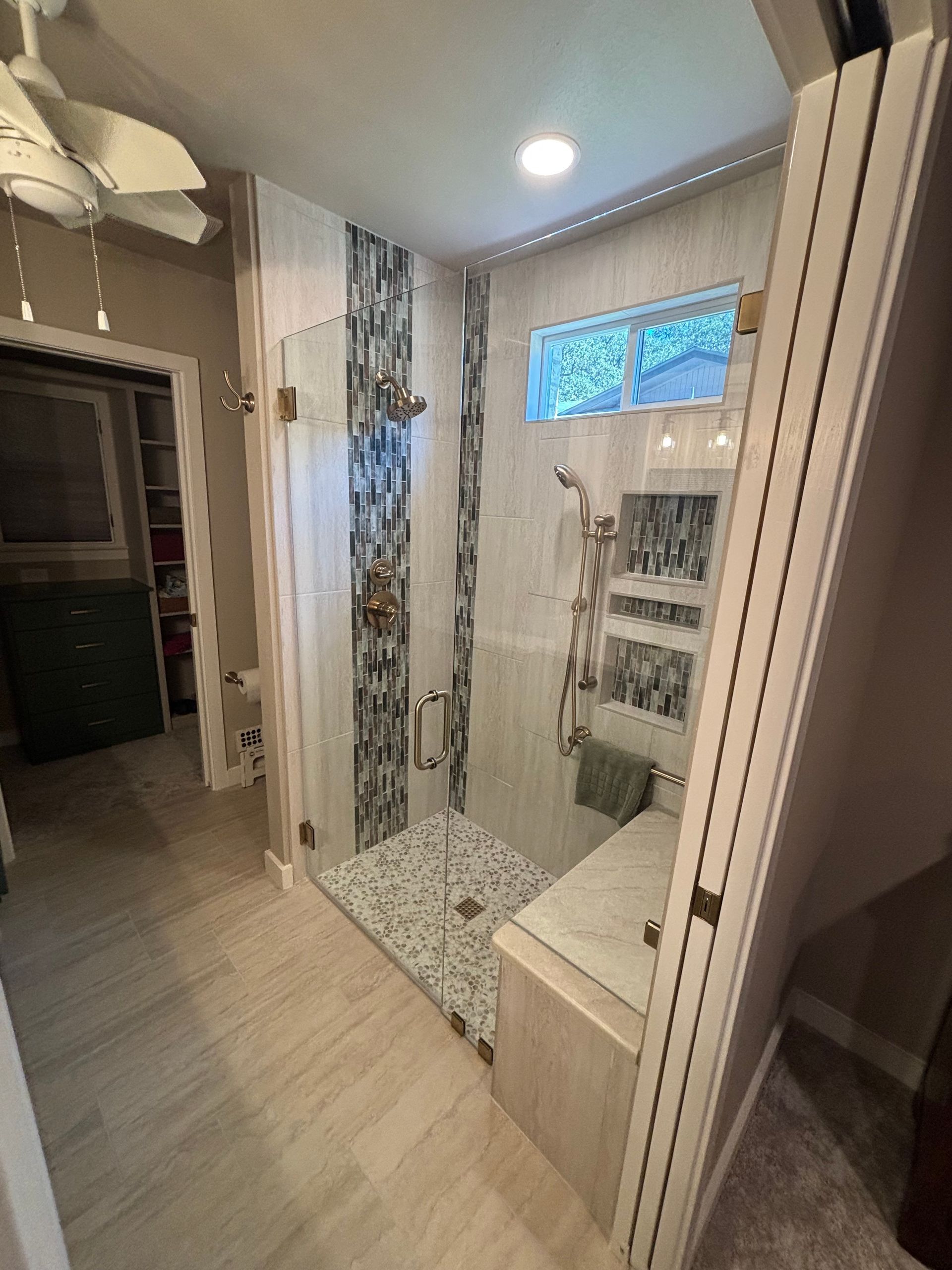 Modern bathroom with tiled shower, glass door, bench, and window. Light wood and blue tile accents.