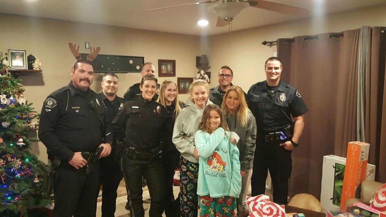 Police officers and several girls smile for a photo with a decorated Christmas tree and presents in a home.
