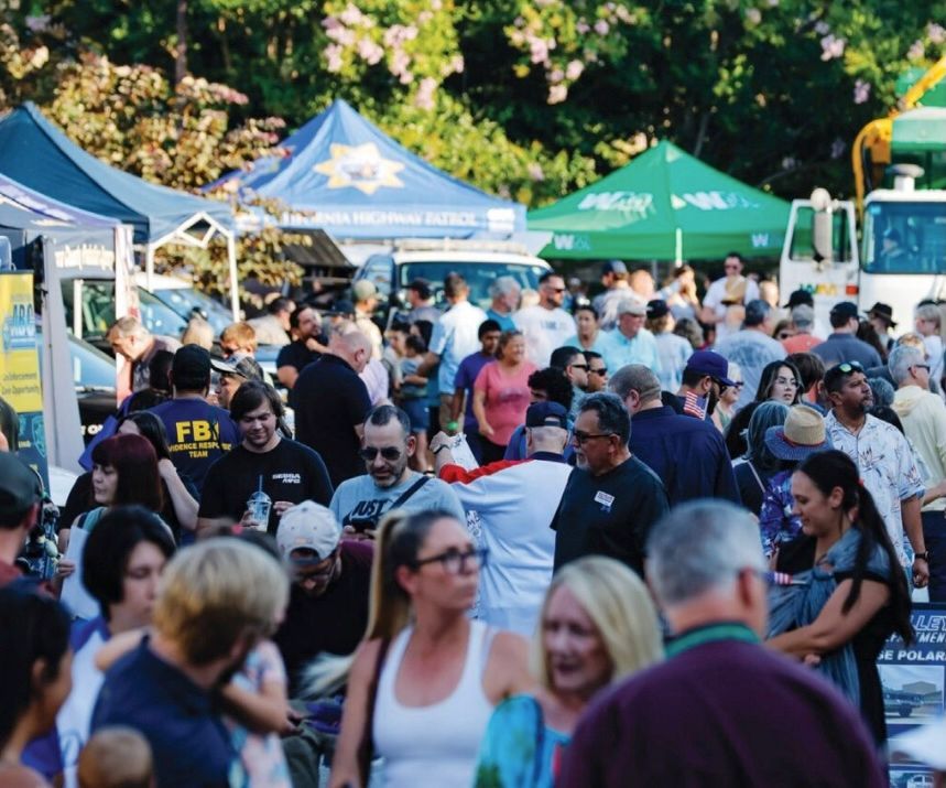 Crowd at outdoor event with vendor tents. Some wear sunglasses and hats; others walk and talk.