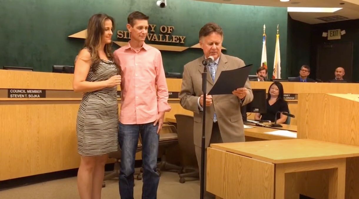 Couple and speaker at a podium in a government setting; man reads document; audience in background.