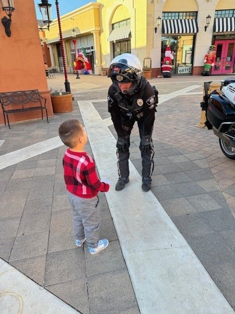 Child in red plaid shirt facing a police officer in black uniform and helmet. They are outdoors.