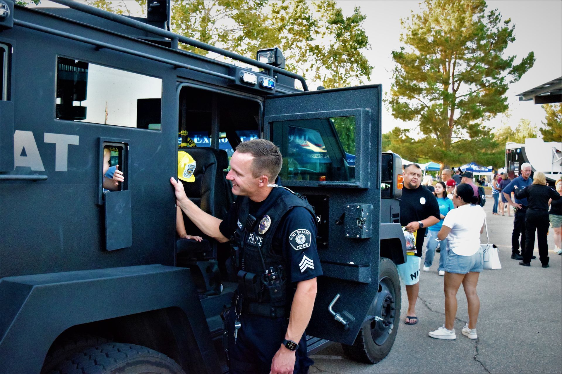 Police officer smiles next to an armored SWAT vehicle; community event with onlookers.