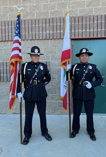 Two uniformed police officers holding American and California flags standing in front of a building.