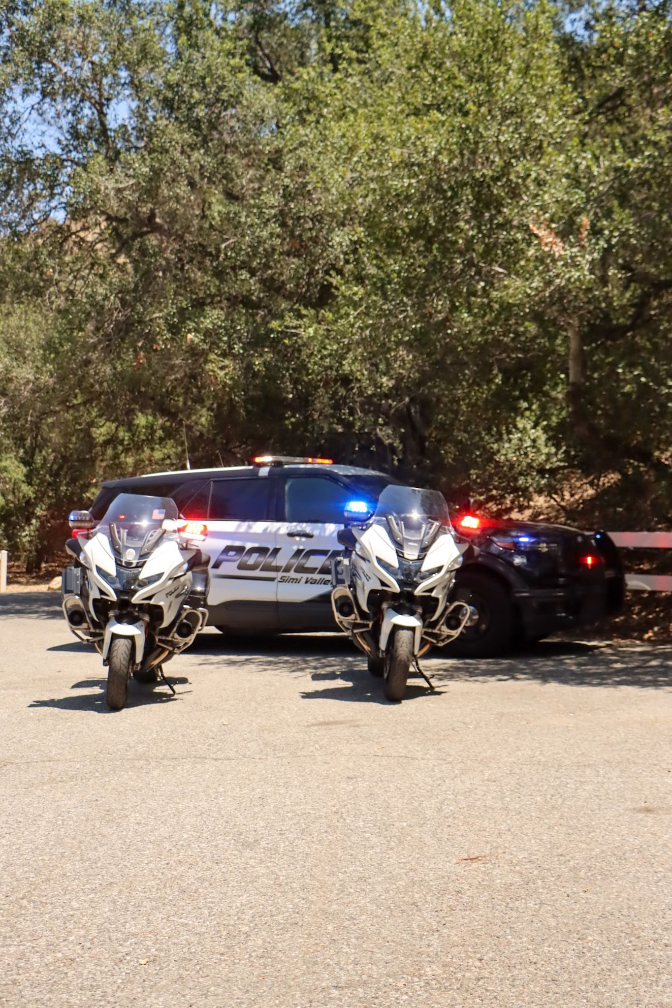 Two police motorcycles flank a police SUV. Bright sunlight shines on the vehicles.
