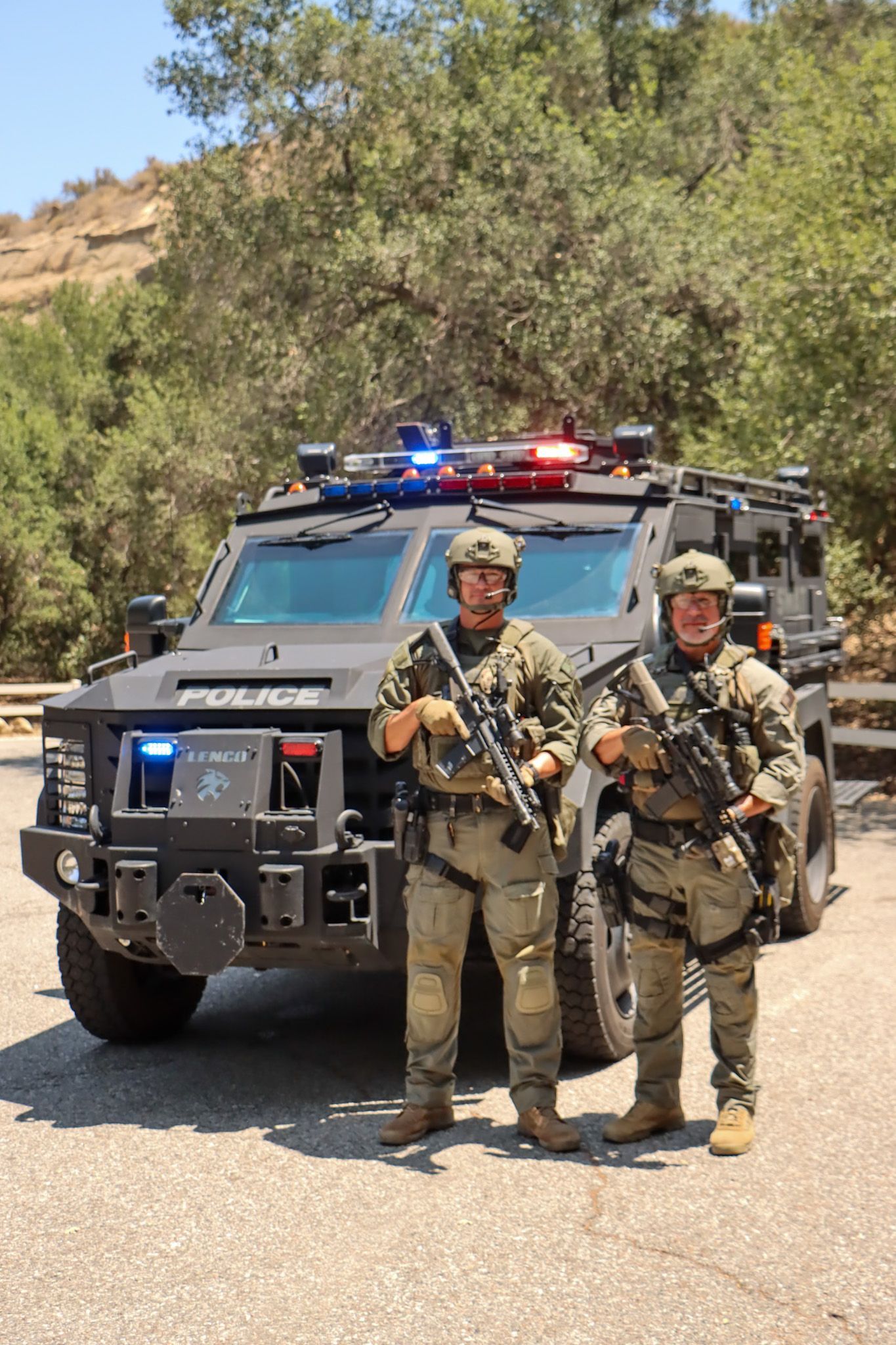 Two armed police officers stand in front of a tactical vehicle in a wooded area.