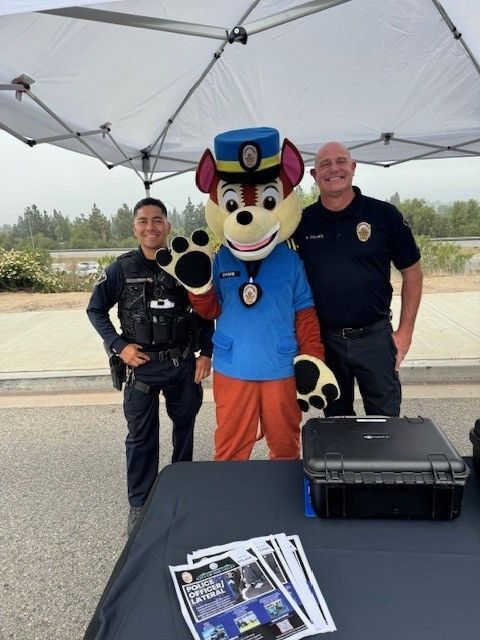 Police officers and mascot at a community event. Under a canopy, officers pose with the mascot and table of materials.