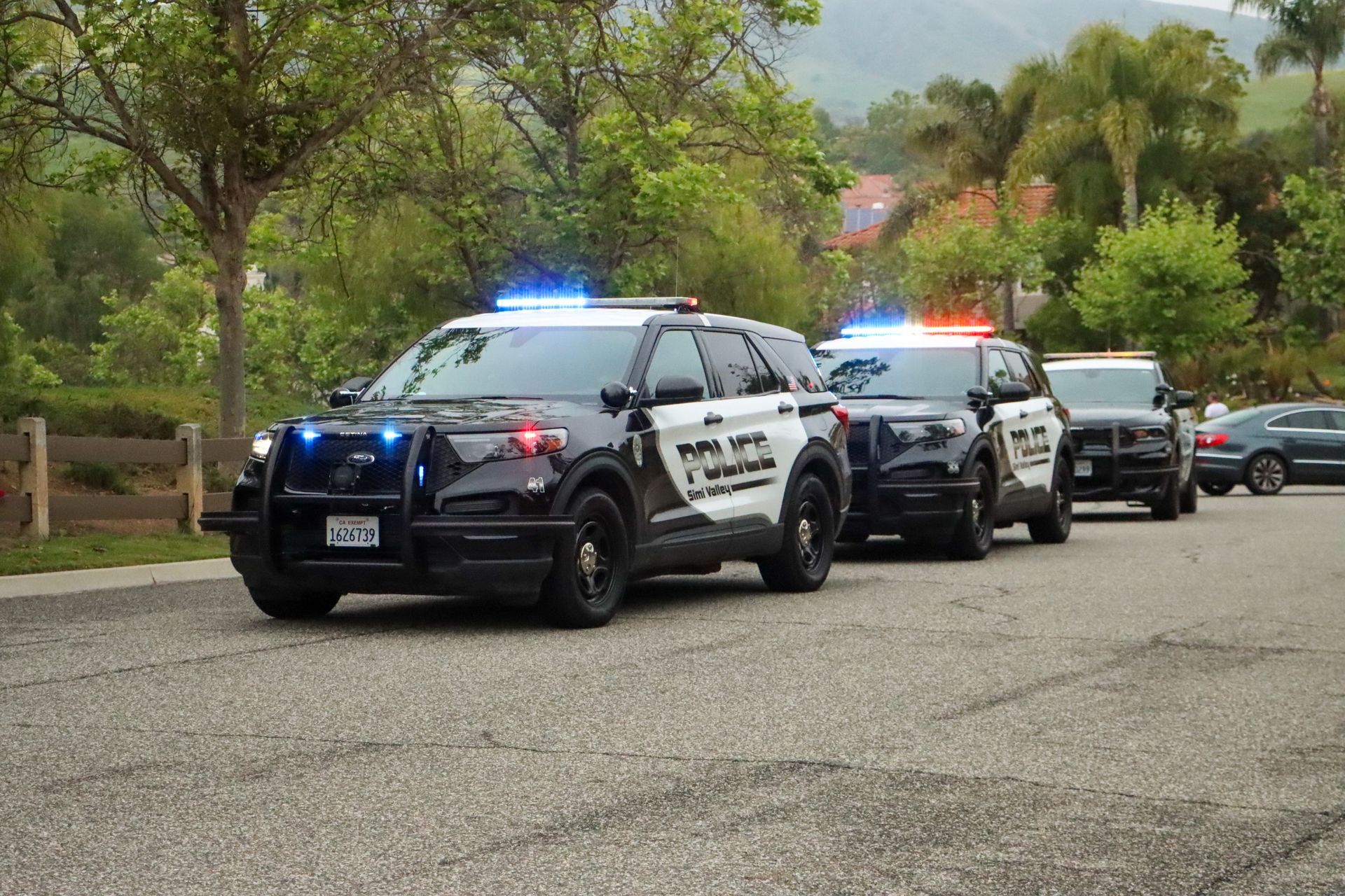 Police SUVs with flashing lights parked on asphalt, near trees and a hill.