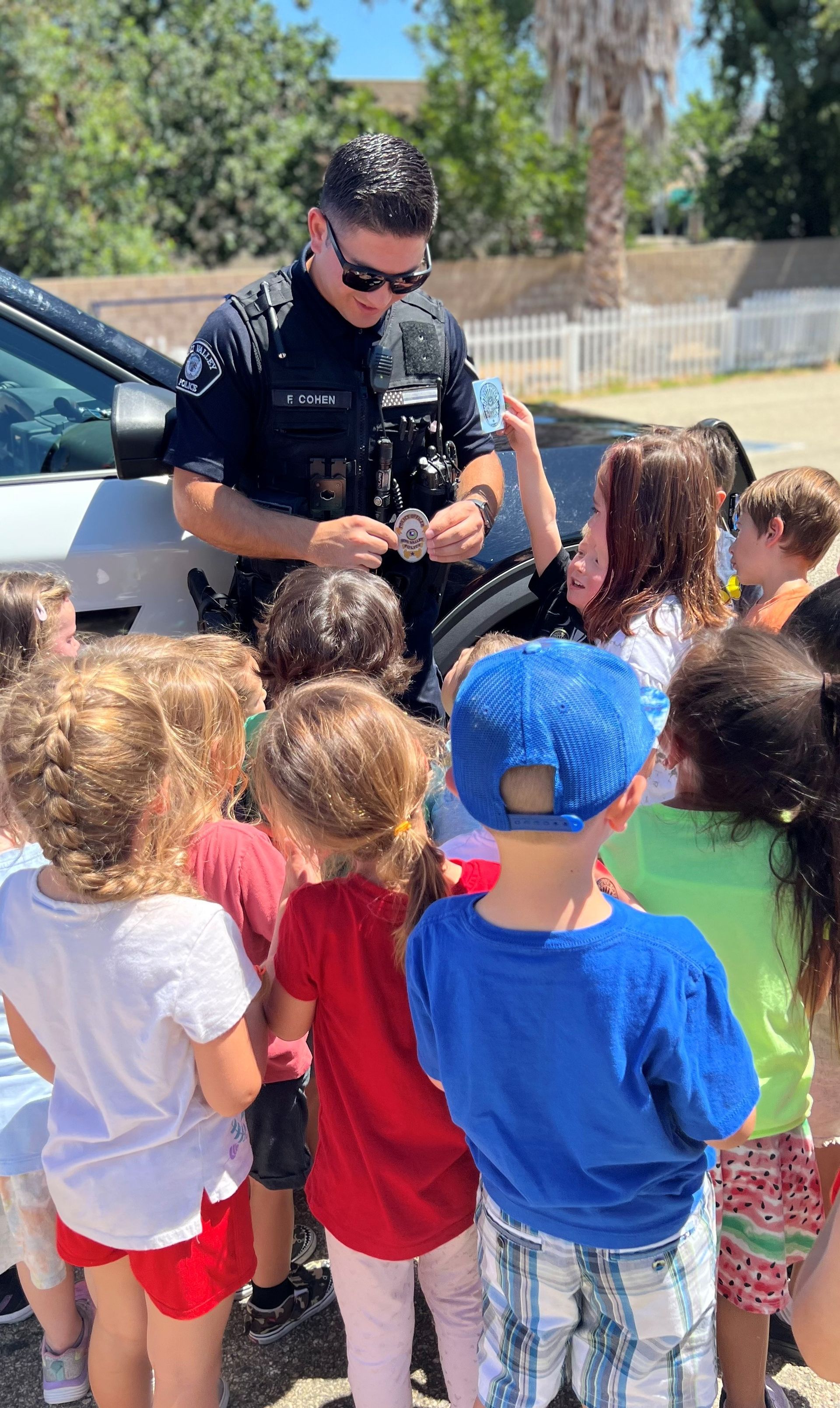 Police officer shows badge to a group of children near a patrol car outdoors.