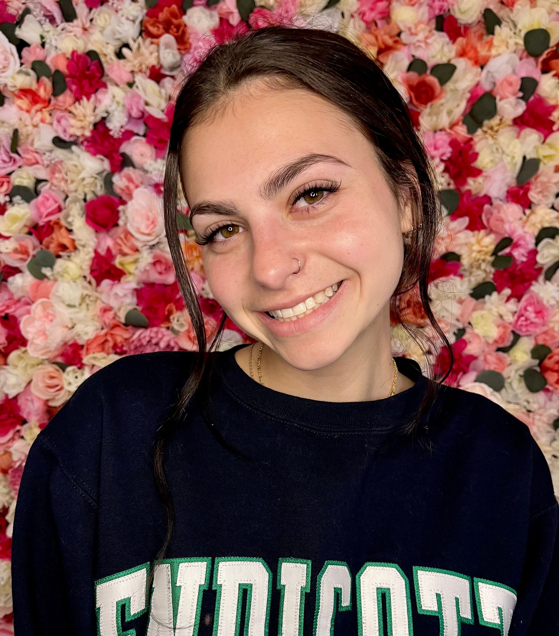 A young woman wearing a sweater is smiling in front of a wall of flowers.