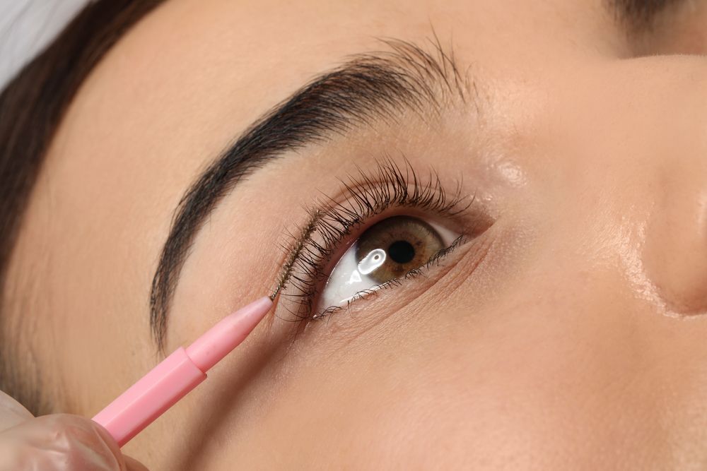 A woman is applying eyeliner to her eye with a pink brush.