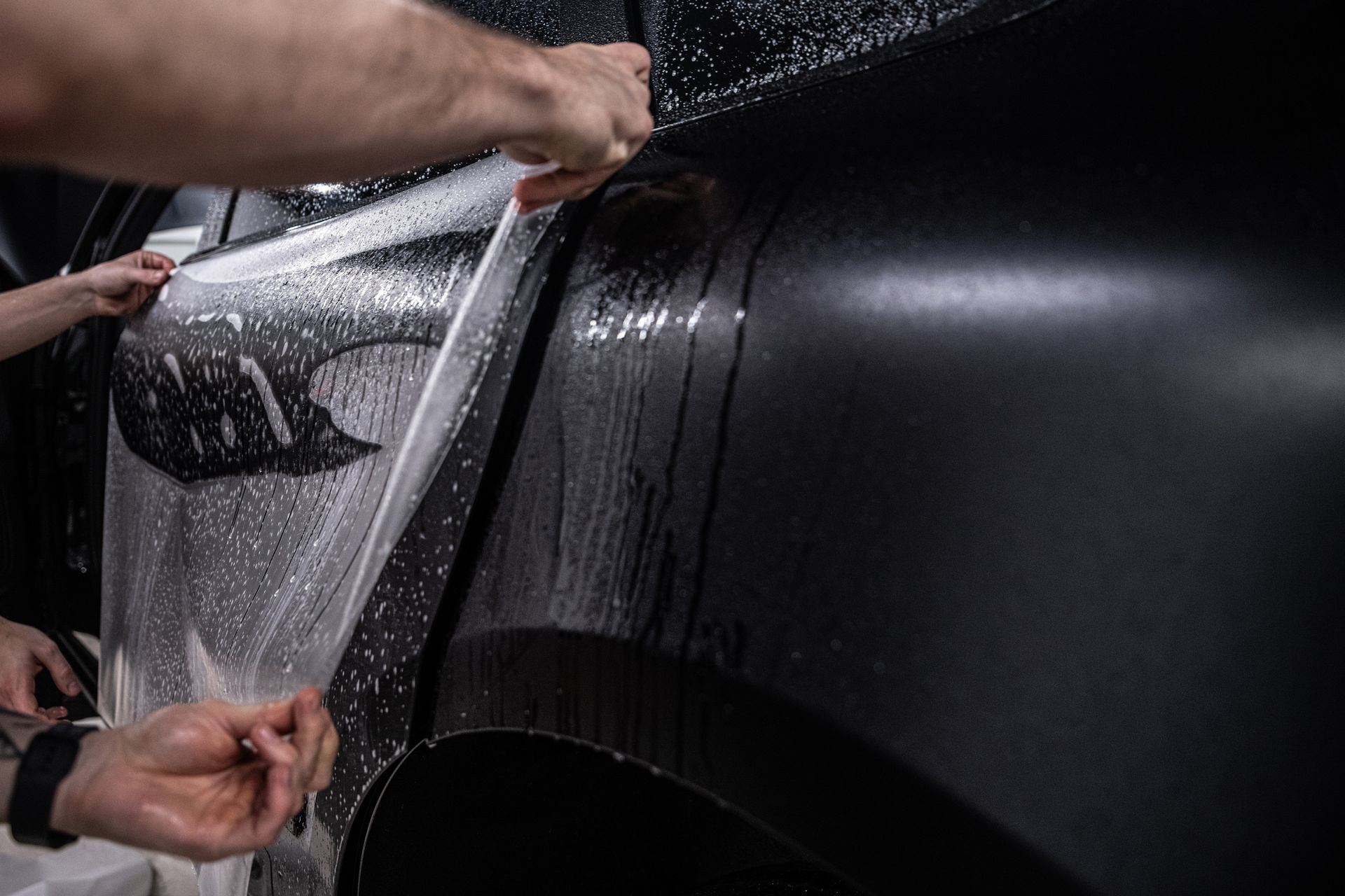 Hands applying a clear vinyl wrap to a black car door, with water droplets visible on the surface.