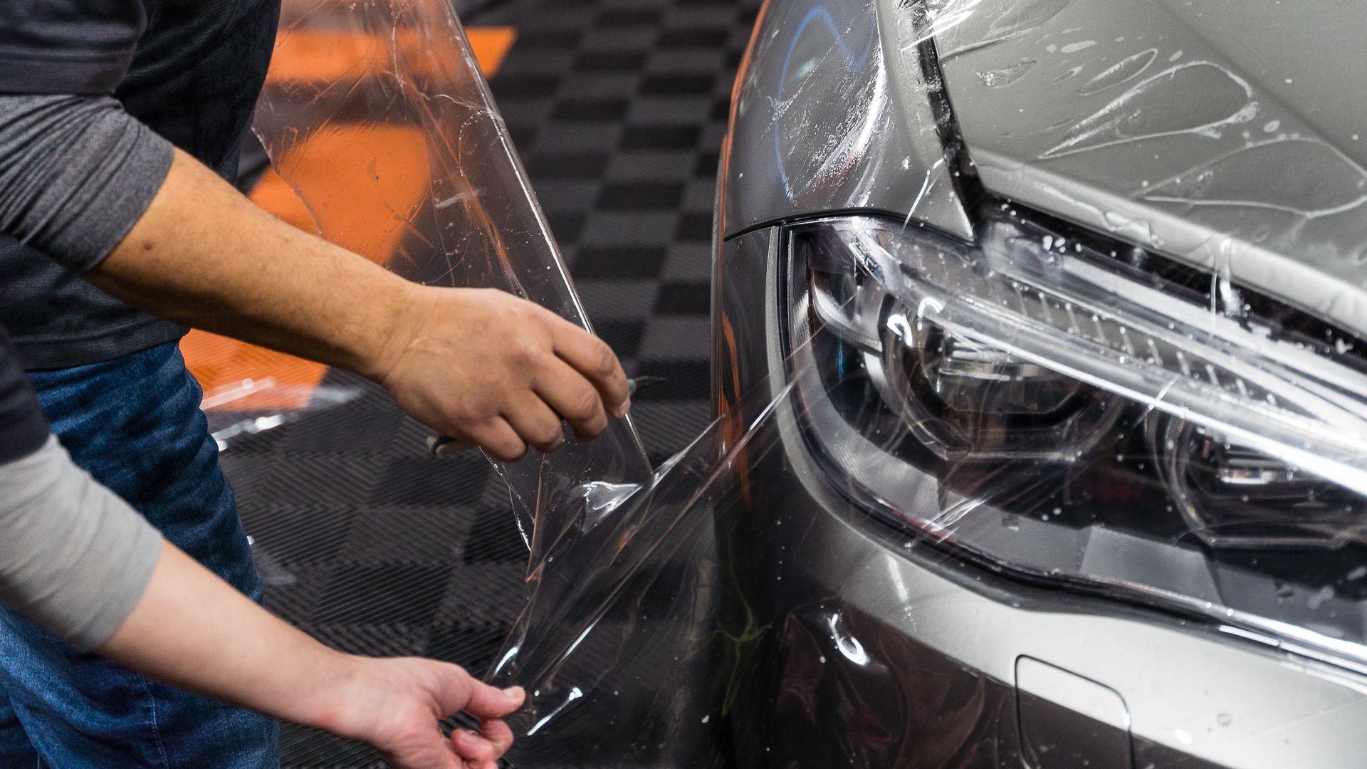 Person applying clear protective film to a silver car front bumper in a garage
