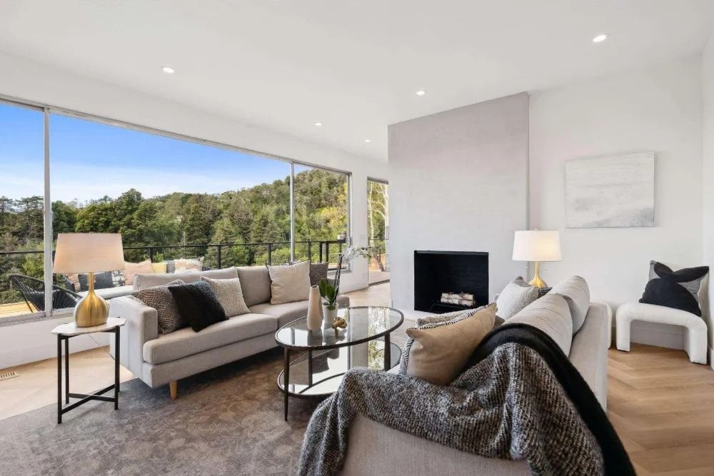Living room with large window overlooking a wooded landscape, neutral-toned furniture, fireplace, and modern decor.