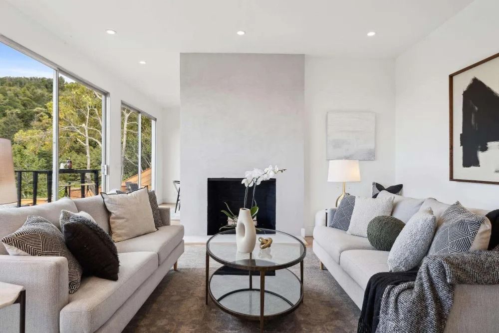 Living room with two white sofas, fireplace, glass coffee table, and artwork overlooking a forest view.