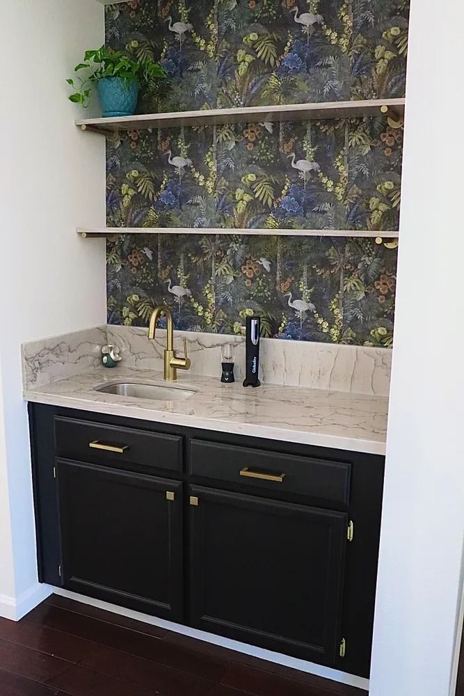 Wet bar with black cabinets, marble countertop, gold faucet, shelves, and patterned wallpaper.