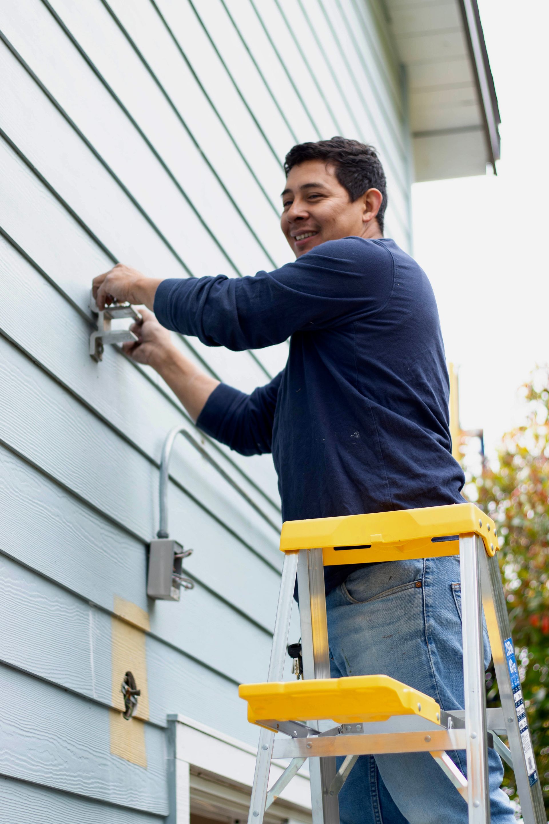 Man on ladder installing light fixture on a light blue siding wall.