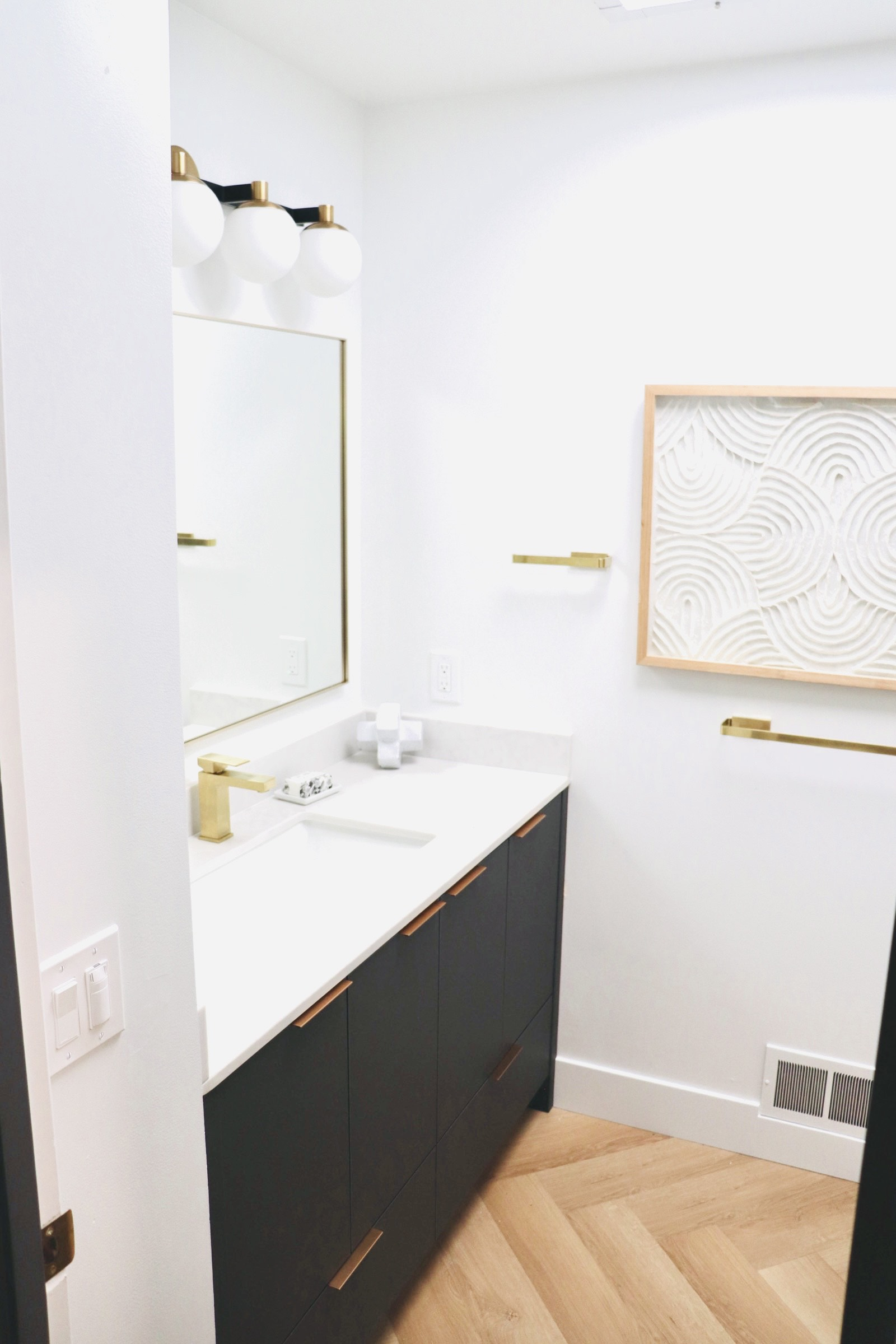 A modern bathroom with black vanity, gold fixtures, white countertop, and artwork on a white wall.