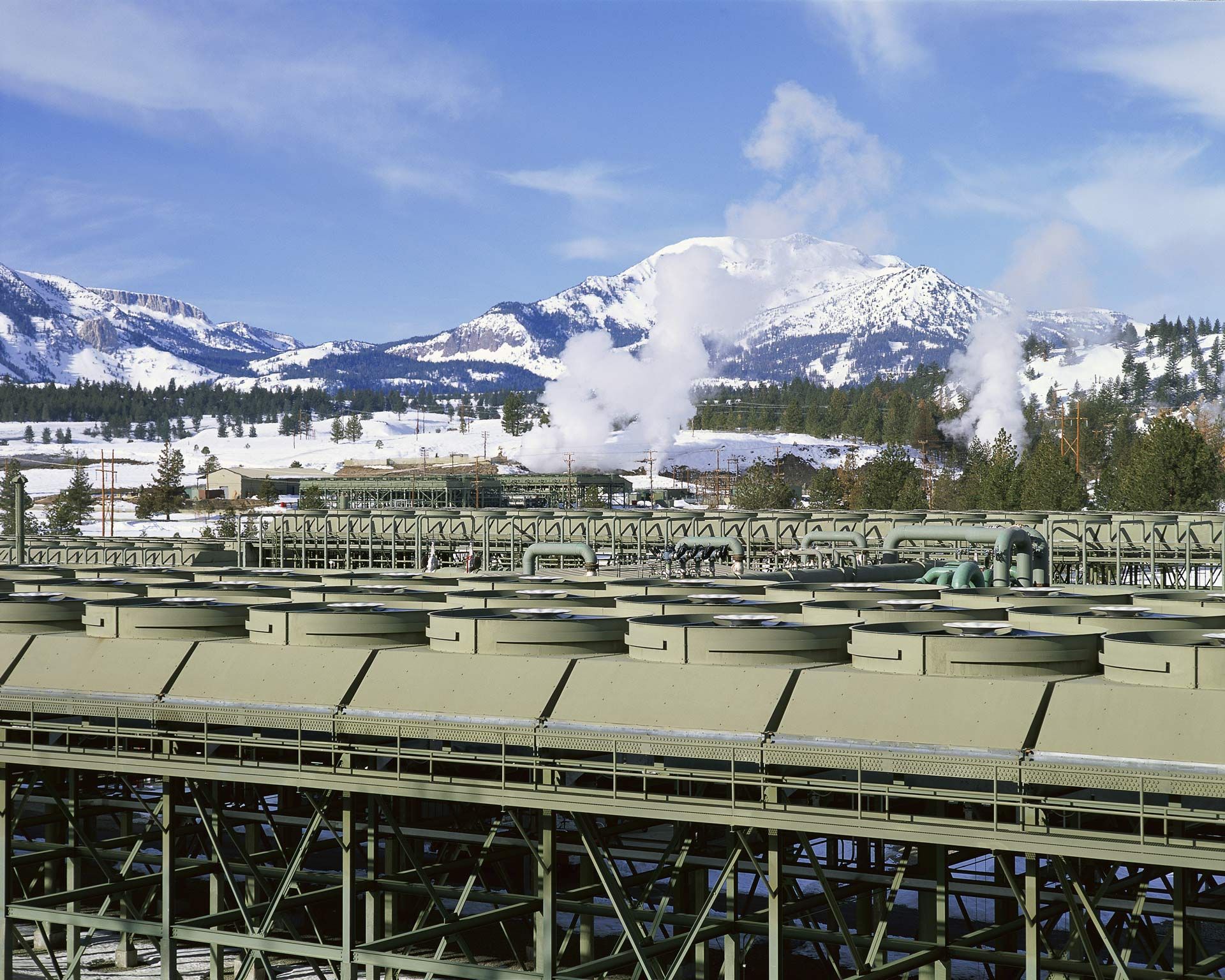 Cooling towers and infrastructure with snowy mountain backdrop, steam rising.