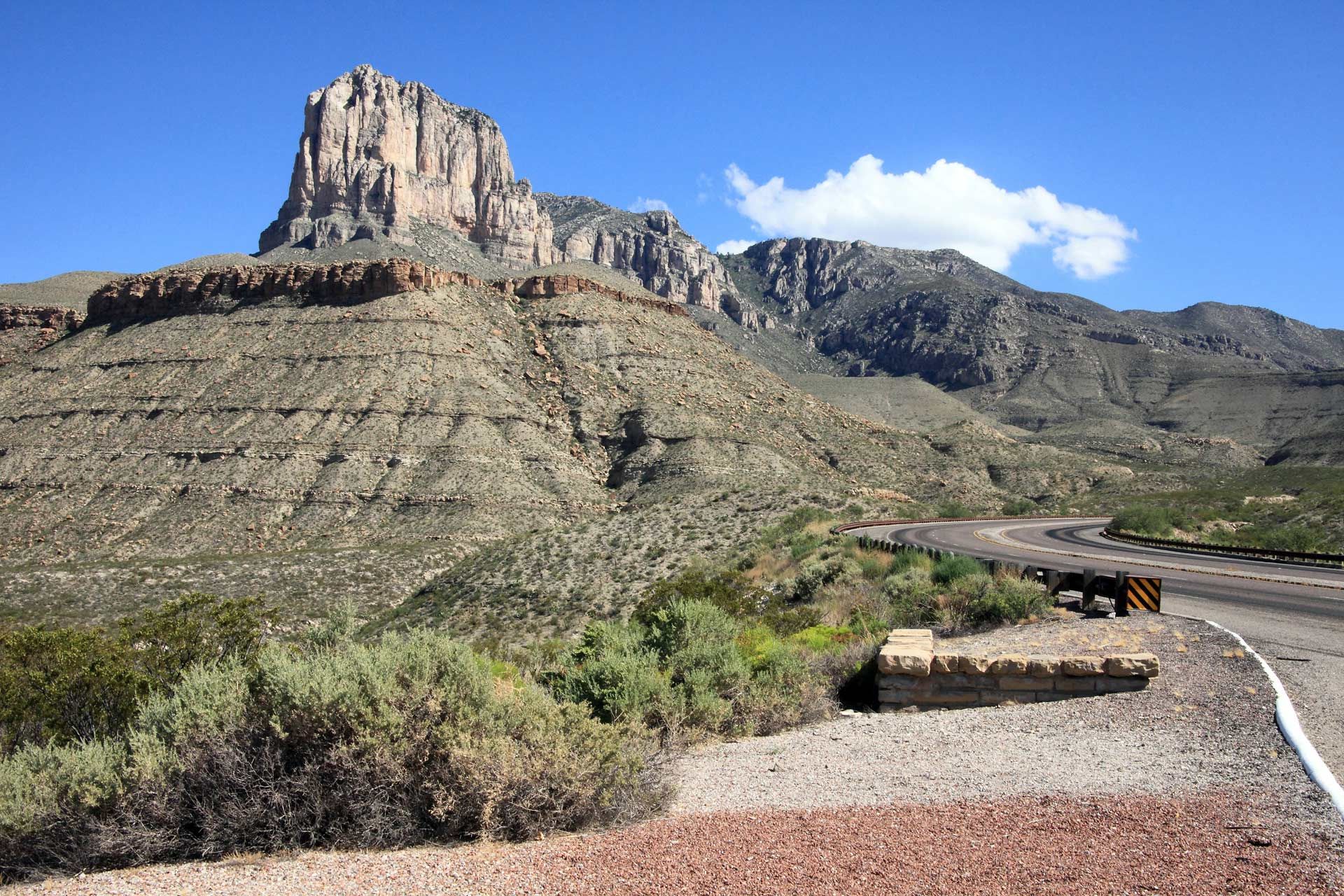 Rocky mountain with a road in the foreground under a blue sky, some greenery.