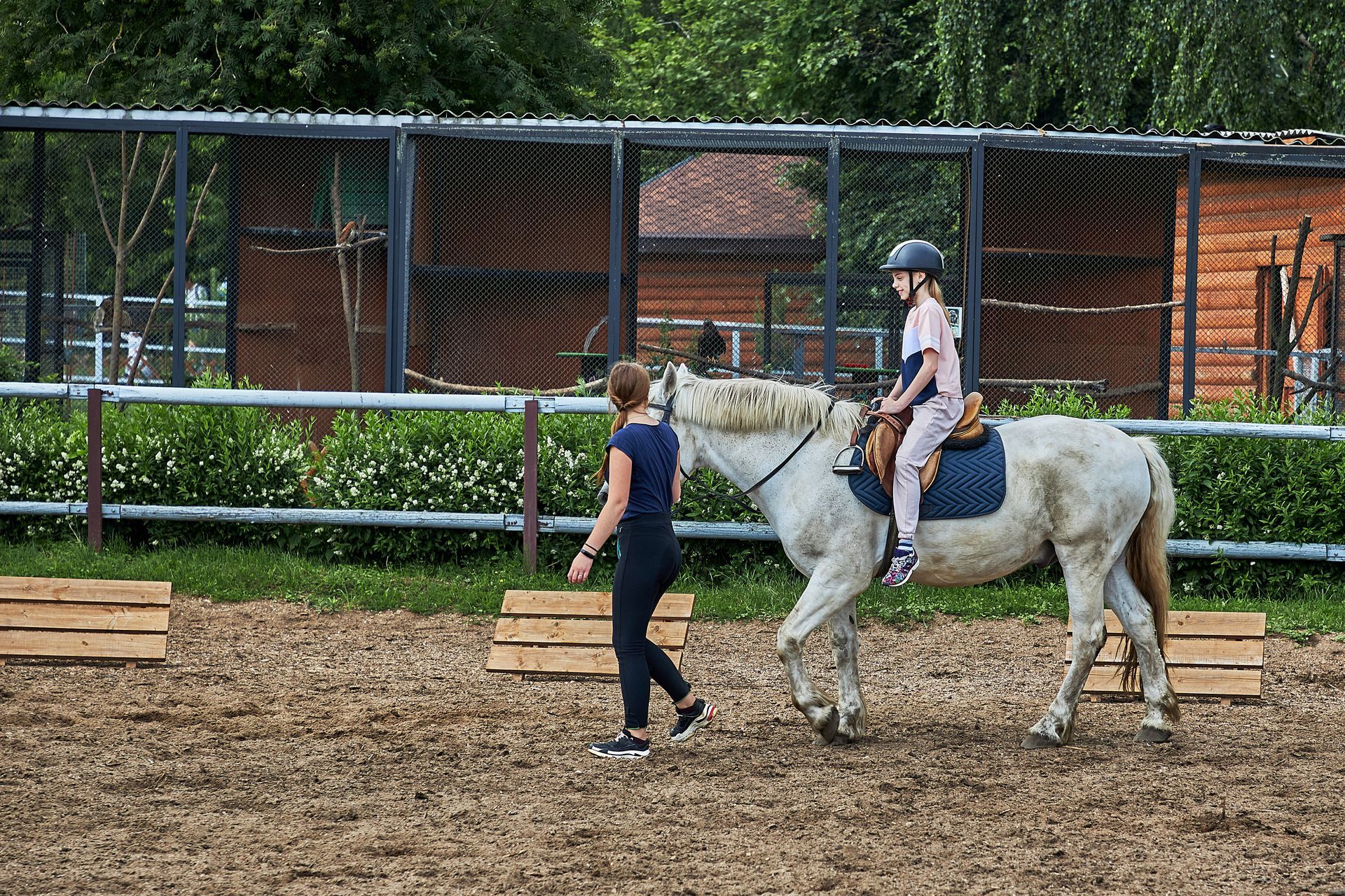 A woman is walking a child on a horse in a fenced in area.