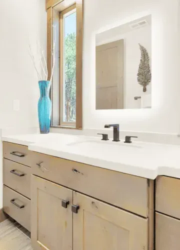 Bathroom vanity with light-up mirror, blue vase, wooden cabinets, and a window with a view by Syn-Mar